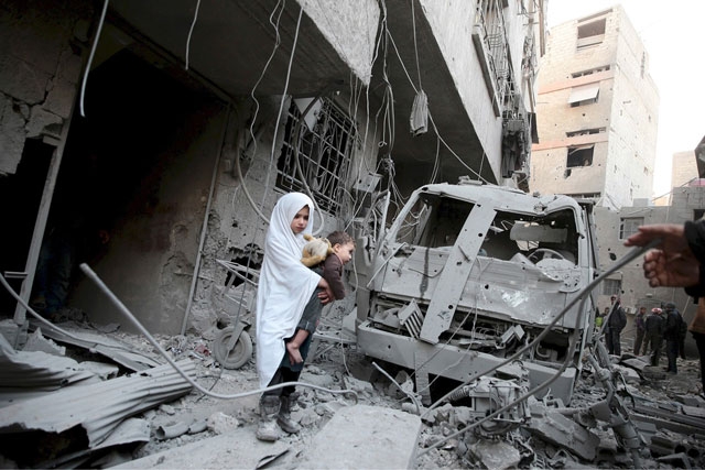 فتاة تحمل طفلاً تراقب مبنى تعرض للدمار جرّاء القتال الذي شهدته منطقة دوما السورية - REUTERS